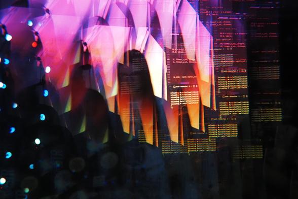 A person doing live coding at their laptops with a projection behind them. The image is distorted slightly due to the use of a kaleidoscopic prism