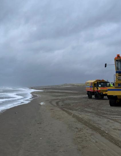 Grauer Himmel, dunkler Sand, links Meer mit flachen Wellen, rechts große Fahrzeuge der Seenotrettung