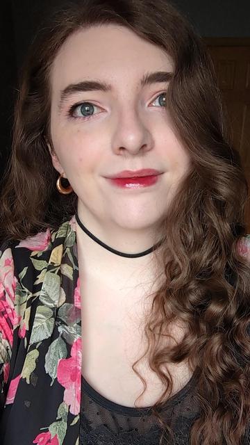 Tulip smiling at the camera. they're wearing a vibrant red-pink lipgloss, rose gold hoop earrings, a black and pink floral shawl, and a black choker.