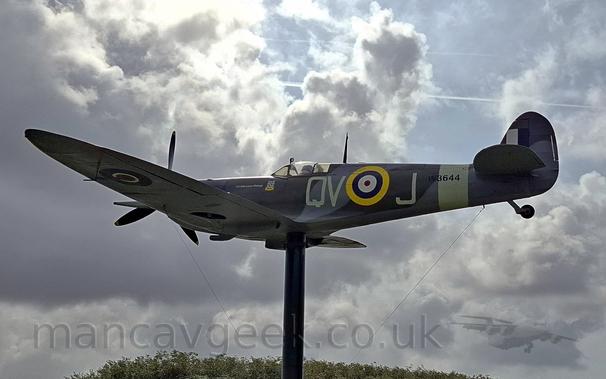 Low side view of a World War 2-era single-engined military fighter pale mounted on a pole in a flying pose.
The plane is in a grey and green camouflage scheme, with the cream-coloured letters "QV-J" either side of a yellow, blue, white, and red roundel on the fuselage.
There is a cream band around the rear fuselage, with "W3644" overlaid in black.
The underside of the body and wings are a pale creamy white, with a blue, white, and red roundel on the outer panels of the wings.
The black pole it sits on seems to rise out of the top of a stand of trees, with dramatically lit grey clouds and blue sky above.