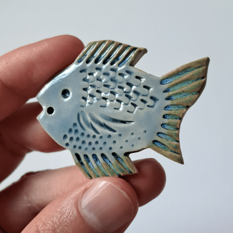 A hand holding a stoneware clay fish pendant. It's a simple fish design, flat, with patterns squished in. It's coloured with blue, transparent glaze on the body and a shifting blue-green glaze on the fins.