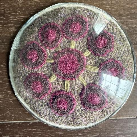 A head-on view of a frameless convex circle of glass over an embroidery of an abstract flower comprised of eight pink circles with deep purple layers surrounding a similar larger circle in the center. The outer circles are connected to the center with green stems. The flower is made with French knots on a background of green and purple variegated cross stitches.