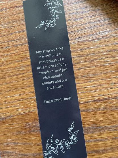 A black bookmark on a wooden table. In white outline is flowering vine on top and bottom edges. In center is Thich Nhat Hanh quote:

Any step we take 
in mindfulness
that brings us a 
little more solidity,
freedom, and joy
also benefits
society and our
ancestors.