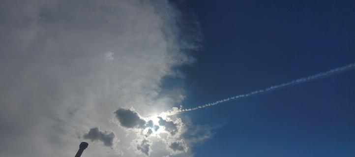 A condensation trail from an airplane points straight at the sun behind a few small clouds, looking like a bright explosion.