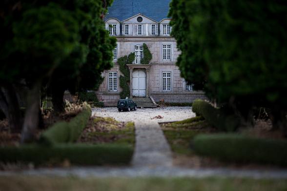 Miniature of a classic-style mansion with a central doorway, flanked by trees and topiary, with a toy car on the driveway.