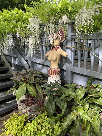 Ceramic sculpture of a stylized female figure with truncated arms and a kind of Greek looking skirt and a massive stylized headdress that looks like it might be an alien or an African mask. It's buff colored, but glazed ceramic with red, white, and black under glaze added in some areas. It is flanked by several large ceramic urns containing exotic looking ferns and small rhododendrons and chartreuse leaved creeping Jenny plants