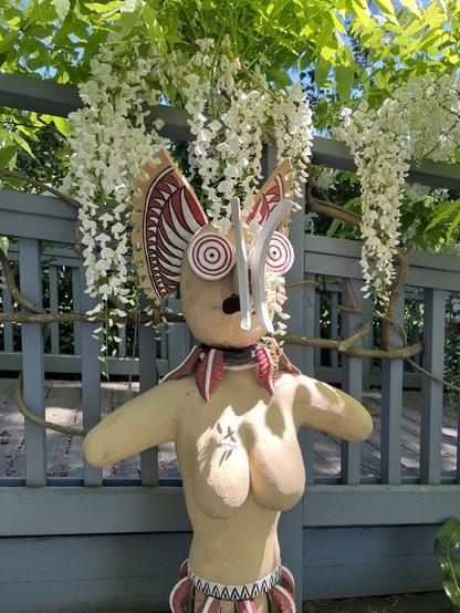 Closer view of the head of the sculpture. The eyes look like round discs or radio telescope discs. The headdress is red and white flares like wings. A pair of nasal tusk like ornaments protrudes from where her nose should be. Her mouth is open in an expression of dismay.