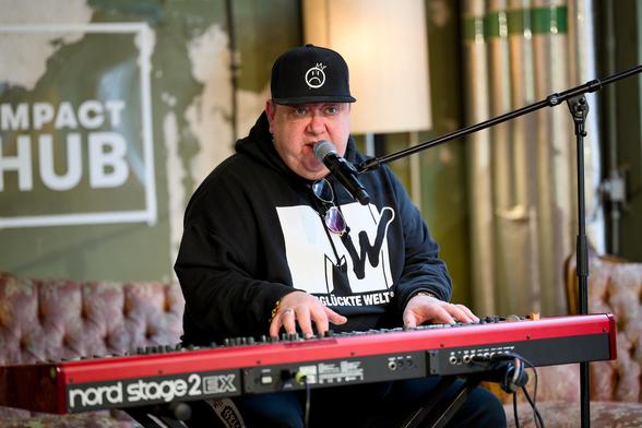 Ein Sänger mit schwarzem Hoodie und schwarzer Cap sitzt an einem roten Keyboard und singt in ein Mikrofon. Auf seinem Hoodie ist ein Aufdruck, der dem MTV-Logo ähnelt. Darunter steht "Missglückte Welt".