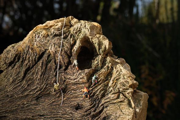 The image features a rugged rock formation with miniature climbers scaling its surface. A climbing rope extends from the top, and a cave entrance is visible on the side of the rock. The rock resembles a goosefish.