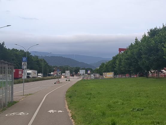 Fahrrad und Fußweg im Bildvordergrund mittig. Rechts: Wiese und Bäume.

Blick auf wolkenverhangene Hügel im Bildhintergrund