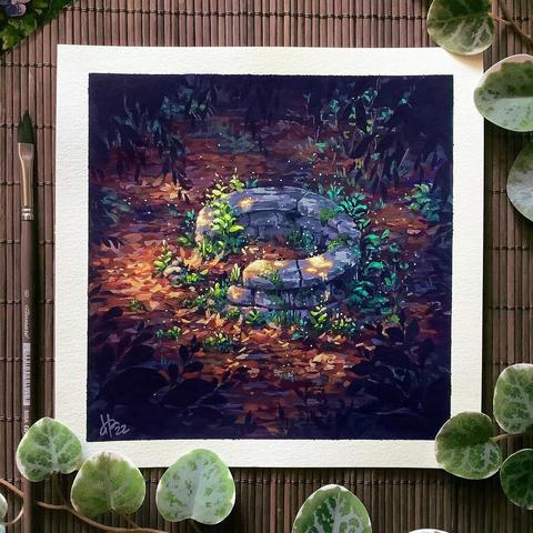 A photo of a square painting placed on a dark wooden mat and surrounded by a paint brush and leaves for decoration. The painting features the remains of a round stone well in the forest, its interior and surroundings filled with warm-coloured autumn foliage. Green plants grow around the well’s rim and soft dappled light falls on it. Further from the well the forest fades into shadow.