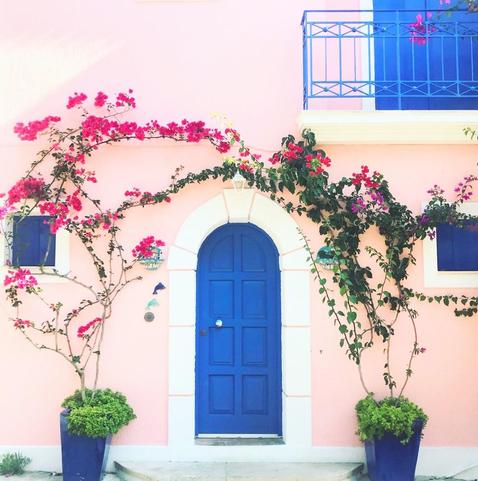 Beautiful rounded blue door way on light painted house with 2 potted plants on either side of the door with the pink plants both climbing like vines above the door way!