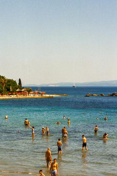 The image captures a vibrant beach scene. Several people are enjoying the cool water on a bright sunny day. In the background, beach umbrellas and sunbeds line the sandy shore of Split, creating a relaxing vacation atmosphere. Further out, a sailboat glides along the tranquil waters, enhancing the picturesque view.