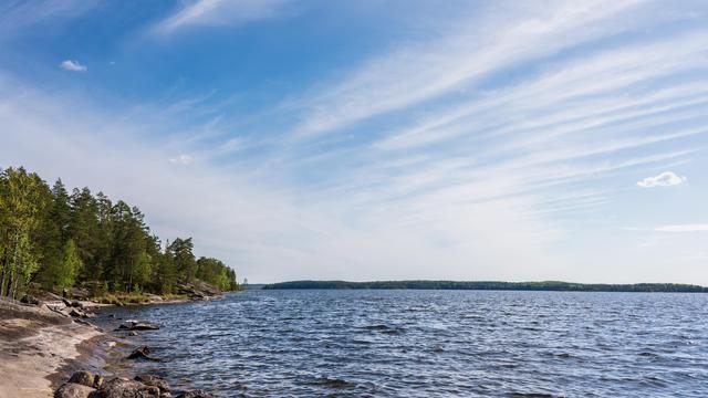 Lake with rippling water under a blue sky filled with wispy clouds. Dense row of trees along the rocky shoreline on the left. Distant landmass visible on the horizon, partially covered with greenery.