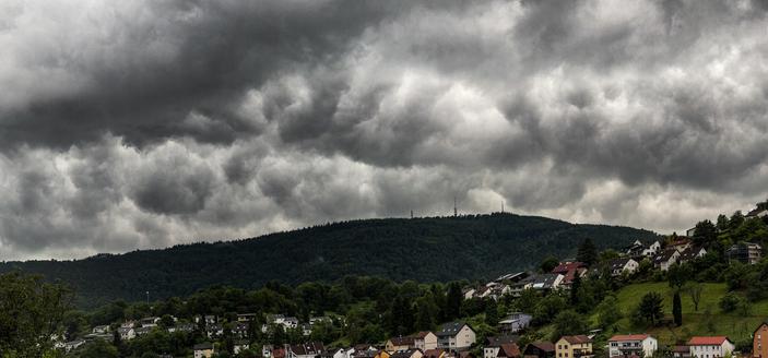 A line of dark grey clouds seen above a large green hill, with some houses in the foreground.