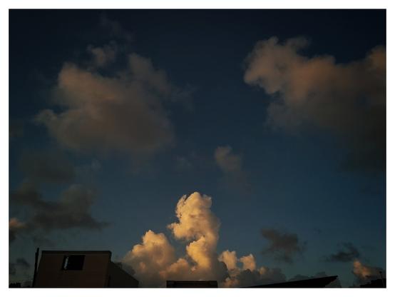 the soft sunlight illuminating the clouds gives them a warm, glowing appearance while the deep blue sky adds contrast. The silhouettes of buildings below add depth and a sense of scale.