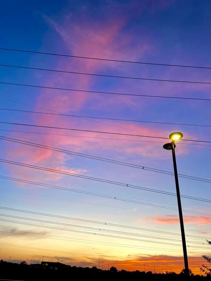 A sunset sky with pink trailing clouds against blue above, and yellow and deep orange on the horizon. The sky is cross crossed with per lines, and a parking lot light stands tall at the right.