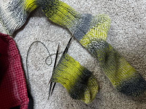 a variegated handknit ribbed wool sock in green and grey. one is complete and the other is knit from the toe to about halfway up the foot.