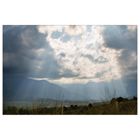 A mountain valley landscape under a dramatic cloudy sky with sunrays breaking through. Tall grass in the foreground adds depth to the wide scenic view.
