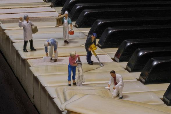 A miniature scene on a piano keyboard depicts small figures engaged in various cleaning and maintenance tasks, such as painting, mopping, and using tools.