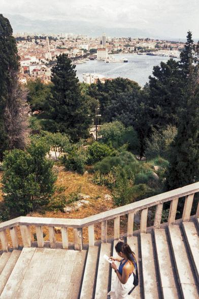 The image captures a lone young adult descending a set of stone stairs overlooking a scenic vista in Split, Croatia. The aged stairs and a weathered stone railing dominate the foreground. Beyond, lush greenery gives way to the Adriatic Sea, with the city of Split nestled along the coastline. The individual appears engrossed in a document or map, seemingly navigating the historical locale.
