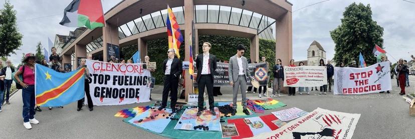Menschen mit Transparenten und Fahnen stehen vor einem Gebäude. Es steht "Glencore Stop Fueling Genocide" auf einem Transparent. Auf einer Welt übersäht mit Kohle stehen drei Anzugträger.