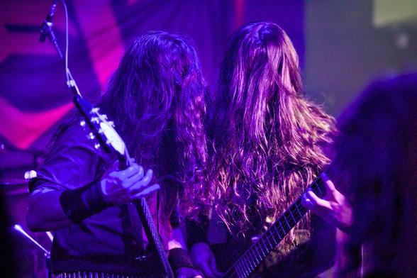 Zwei Personen mit Gitarre und Bass in violettem Licht. Bei beiden verdecken die langen Haare das Gesicht.