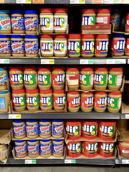 3 rows of grocery store shelves full of peanut butter in jars and on lowest shelf, large canisters. Majority are Jif brands with some brands Skippy.