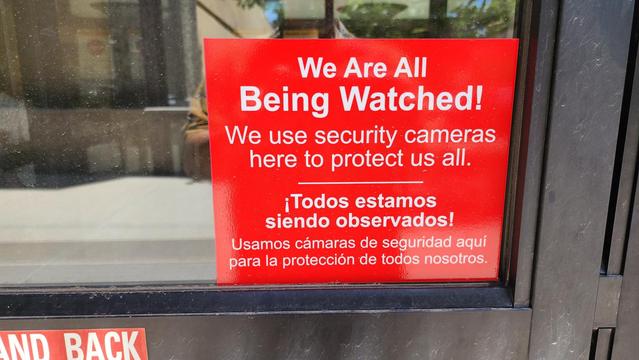 color photo of a red sign with white sans serif that reads, "We Are All Being Watched!" and in Spanish underneath,"Todos estamos siendo observados!"