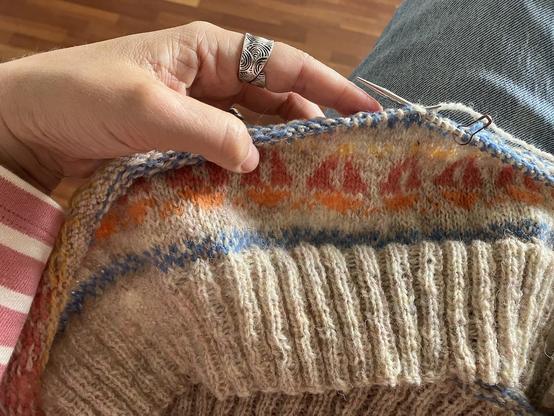 Photo of a knitting project in progress, the pattern is a boat with coral pink sails and an orange hull, the knitter is wearing a pink and white striped jumper.