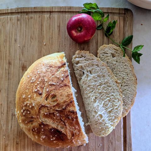 A top-down photo of a loaf of freshly baked bread, sliced and with an apple and mint sprig next to it.