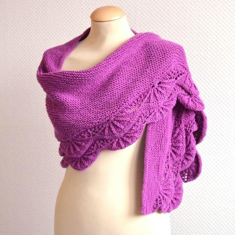 A shawl in pink yarn, with fan-like patterning in the border, and a garter stitch body, is draped over a mannequin against a white background.