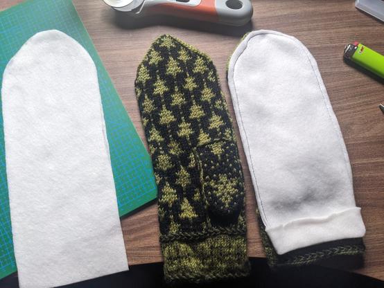 Work in progress on making a thin white fleece lining for knitted gloves. 
Partially on a green cutting mat are two fleece cutouts roughly the size of the knitted mittens. They are knitted in fair isle sporting a repetitive pattern of green pine trees against a black background. Next to the palm-up mitten lies two fleece pieces sewn together atop the other mitten.