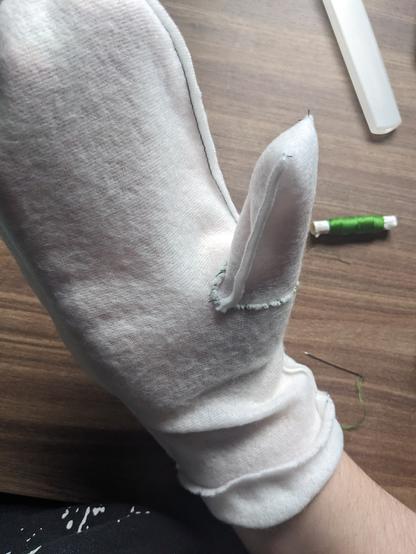 Finished sewn white fleece mitten lining modelled on my hand. The thumb part was whip-stitched with a light green thread. The small spool of which lies on the table in the background.