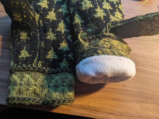 The two finished mittens lying next to each other palms up. The thumb is now standing up a bit more die to the added lining. The right mitten is partially inside out on the cuff part to reveal the whip-stitched white fleece lining.