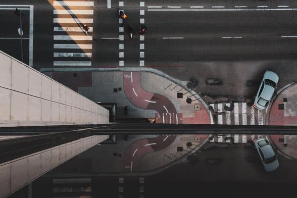 An image showing a street scene from the top of a building, viewed from a top-down perspective. Part of the view is reflected in the building's windows.