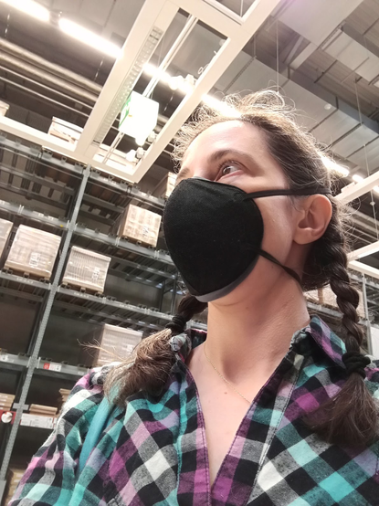 photo of a white woman, her brown and grey hair in two pigtails, is wearing a black respirator and a flannel shirt with a green, purple, black, white, grey check pattern. Behind the woman we see a warehouse with shelves and boxes. The woman is looking off to our left.