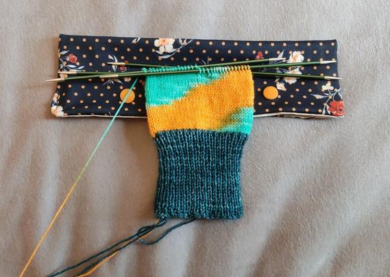 The beginning of a knit sock. The main part is orange and turquoise and the colors are lining up to make a single bold stripe of each color that runs diagonally around the sock. The sock is knit cuff-down, with double-pointed needles, and the cuff is done in a contrasting dark turquoise color. The project is resting on a floral pouch with two snap buttons that hold the needles and make sure the project can't slide off the needles.