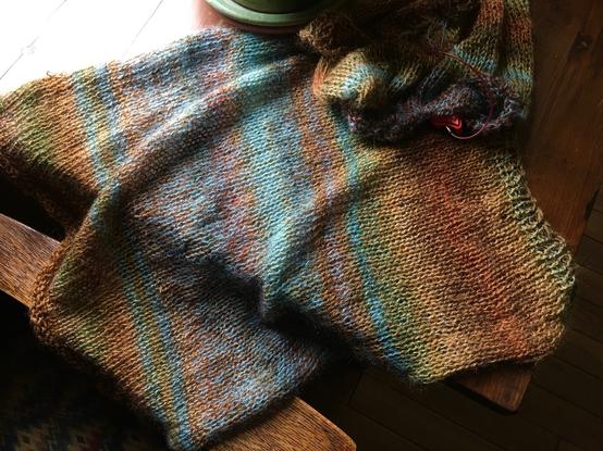 A knitting project: the back of a sweater and the beginning of the sweater front, laid on a table near a window. Colors are varied blended and gradual striping of rusts and blues. The yarn colors slowly change as the pieces are knitted.