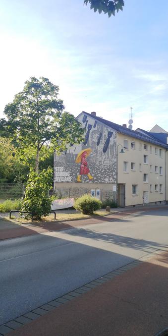 Haus mit Fensterfront zur Straße, die seitliche Hauswand zeigt in einem Wandbild eine gehende Person mit Regenschirm, Regenmantel und Gummistiefeln im Bombenhagel, Beschriftung: Stop bombing civilians