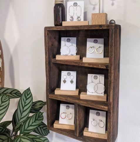 A set of small wooden shelves with hoop earrings of various styles displayed on white cards in wooden holders. The shelves are decorated with other objects such as small brown bottles. Part of a large-leafed variegated plant is in the corner of the image.