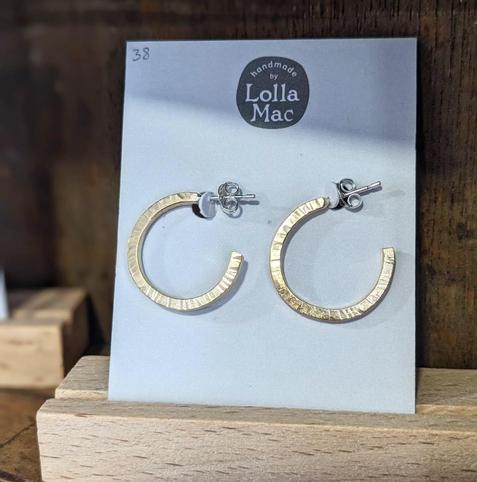 A pair of hoop earrings on a white card displaying the LollaMac logo. The hoops are made of square brass wire formed into a partial hoop, and hammered to create surface texture.