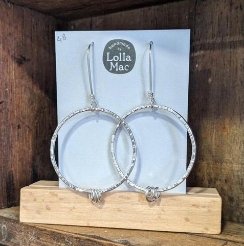 A pair of hoop earrings displayed on a white card bearing the LollaMac logo. The hoops are fine square silver wire, hammered texture, hanging from ear wires, and decorated with small silver rings.