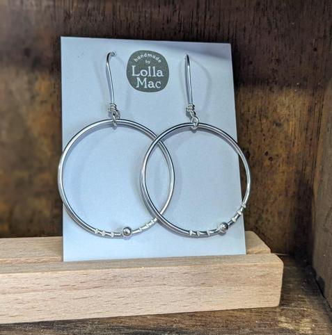 A pair of hoop earrings on a white card bearing the LollaMac logo. The hoops are made of plain round wire, decorated with a single silver ball and just a few hammered indentations, The hoops are suspended from ear wires.