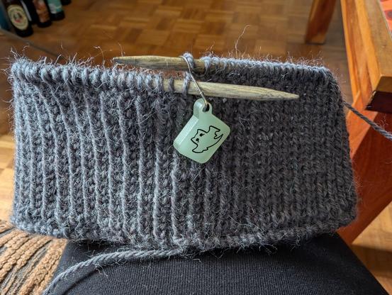 A partially knit hat on wooden circular knitting needles, resting on my thigh. The hat is being knit with grey wool and there's a glow in the dark ghost cat stitch marker hanging between the needles. So far only the ribbing has been knit.