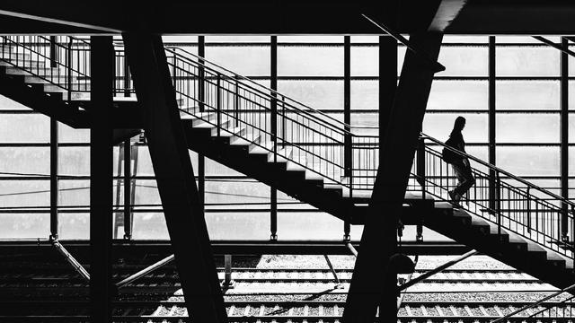 A black and white photo showing a person silhouetted on a staircase inside a modern train station. The scene is framed by angular beams and large window panels that cast a grid of light and shadows over the staircase and train tracks below.