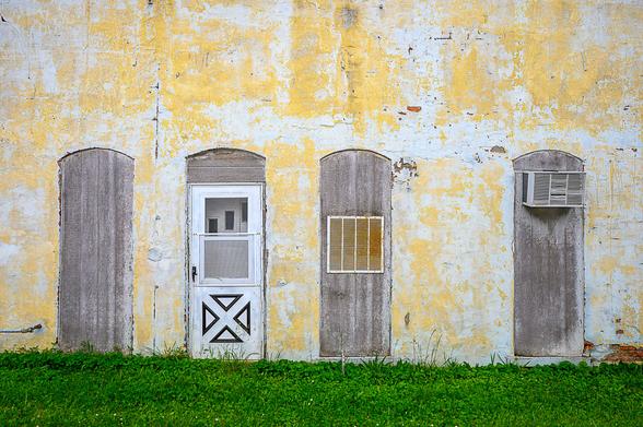 A brick building covered in layers of peeling and fading yellow and white paint.  There are four old openings for doors and/or windows that have been boarded up.  One contains an entrance door, one contains a large vent, and another has a window air conditioner mounted in it.