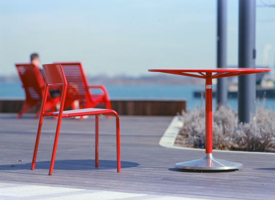A bright red table and chair sit in the boardwalk sun by the lake.