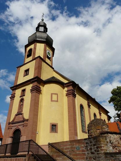 Kirche St. Gertraud Elsenfeld
Die römisch-katholische Kirche St. Gertraud, vollständiger Name St. Ubaldis und Gertrudis, ist ein denkmalgeschütztes Kirchengebäude, das in Elsenfeld steht, einem Markt im Landkreis Miltenberg (Unterfranken, Bayern).
Die Saalkirche, deren Wände außen mit Pilastern gegliedert sind, wurde 1767 gebaut. Das Langhaus aus drei Jochen hat im Osten einen eingezogenen Chor aus einem Joch mit einem dreiseitigen Abschluss. Der im Westen vorgestellte Fassadenturm, der von Treppentürmen flankiert wird, hat ein oktogonales Obergeschoss, das die Turmuhr und den Glockenstuhl beherbergt und auf dem eine Welsche Haube sitzt. Über dem Portal in der Westwand des Turms befindet sich eine Wandnische mit einer Statue des hl. Wendelin. 
(Bild: Wikimedia Commons, 2010, CC-BY-SA 3.0, Manro)