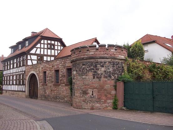 Stadtmauerrest mit rundem Eckturm in Kleinwallstadt an der Ecke Obere Straße - Landesstraße 2309, mit Zinnen, 15./16. Jahrhundert.
Hinten links in Verlängerung des Stadtmauerrestes steht ein Fachwerkhaus.
(Bild: Wikimedia Commons, 2012, CC-BY-SA 3.0, Commander-pirx)